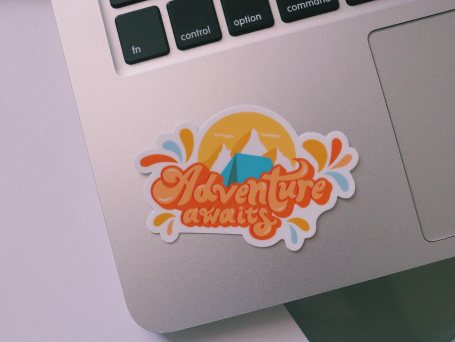 Orange and blue camping sticker that says Adventure Awaits. Includes a mountain range and camping tent
