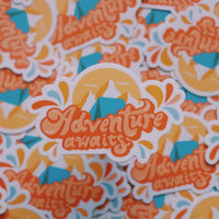 Orange and blue camping sticker that says Adventure Awaits. Includes a mountain range and camping tent