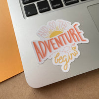 And so the adventure begins glossy vinyl sticker. A hand drawn sticker with sun rising and paper airplane. Sticker is on Macbook laptop bottom left near the keyboard