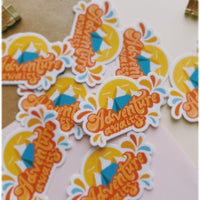 Orange and blue camping sticker that says Adventure Awaits. Includes a mountain range and camping tent