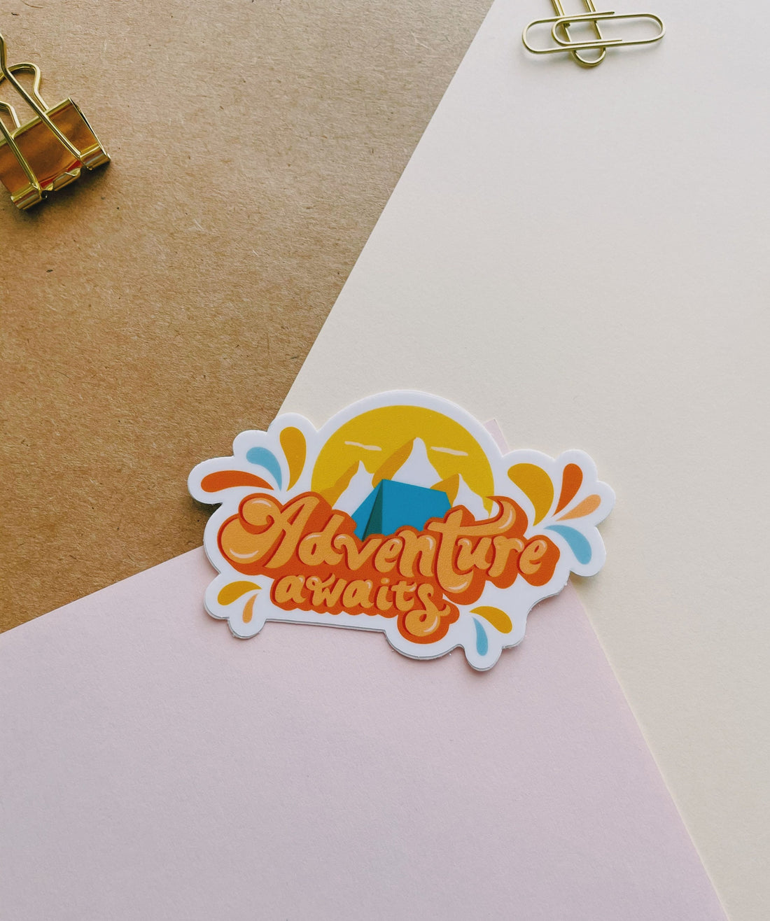 Orange and blue camping sticker that says Adventure Awaits. Includes a mountain range and camping tent