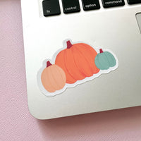 3 pumpkins in one vinyl sticker, one large orange pumpkin in the middle, one medium size cream pumpkin on the left, and one small teal pumpkin on the right. Placed on a Macbook computer near the keyboard