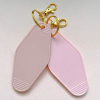 two blank hotel keychains, one beige and one light pink. Both with gold keyring and lobster clasp